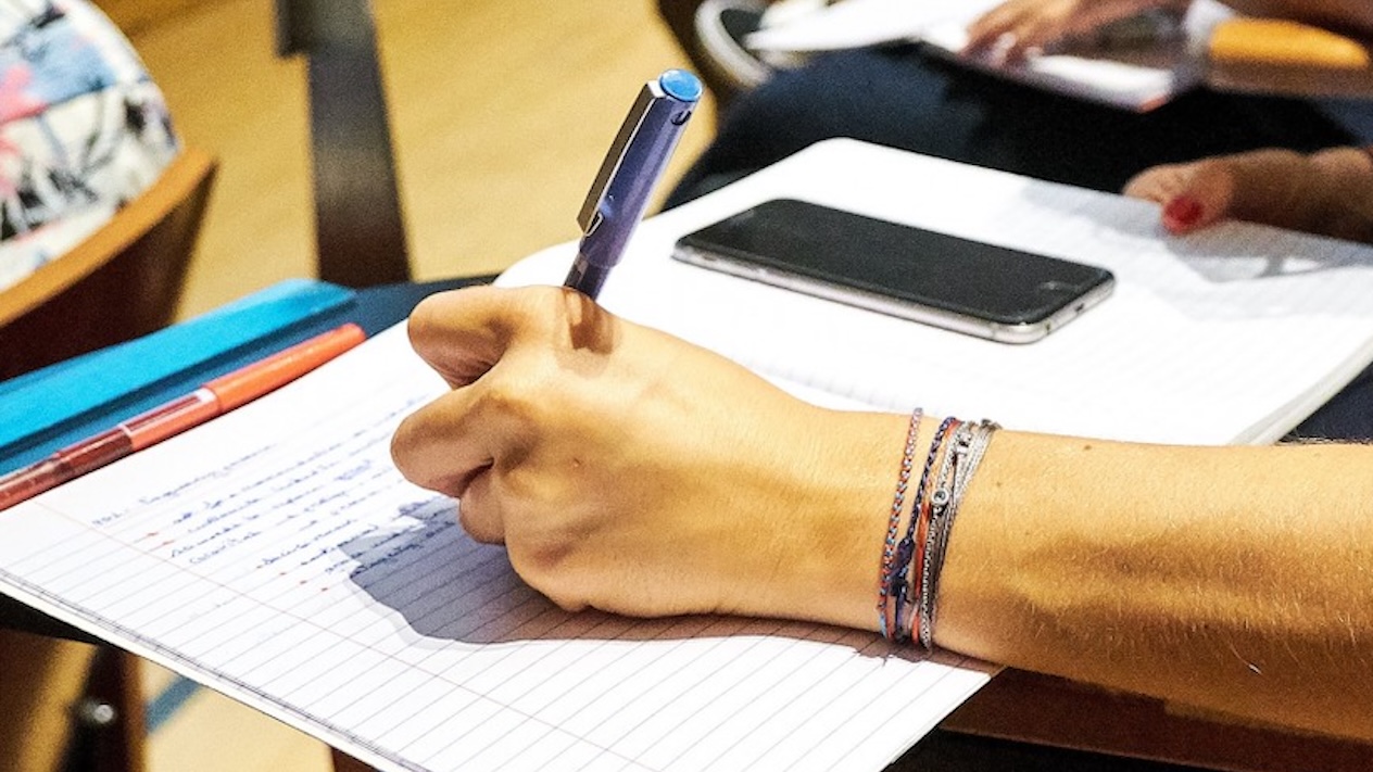 Someone writing notes with a pen during a classroom session, with other participants visible in the blurred background.