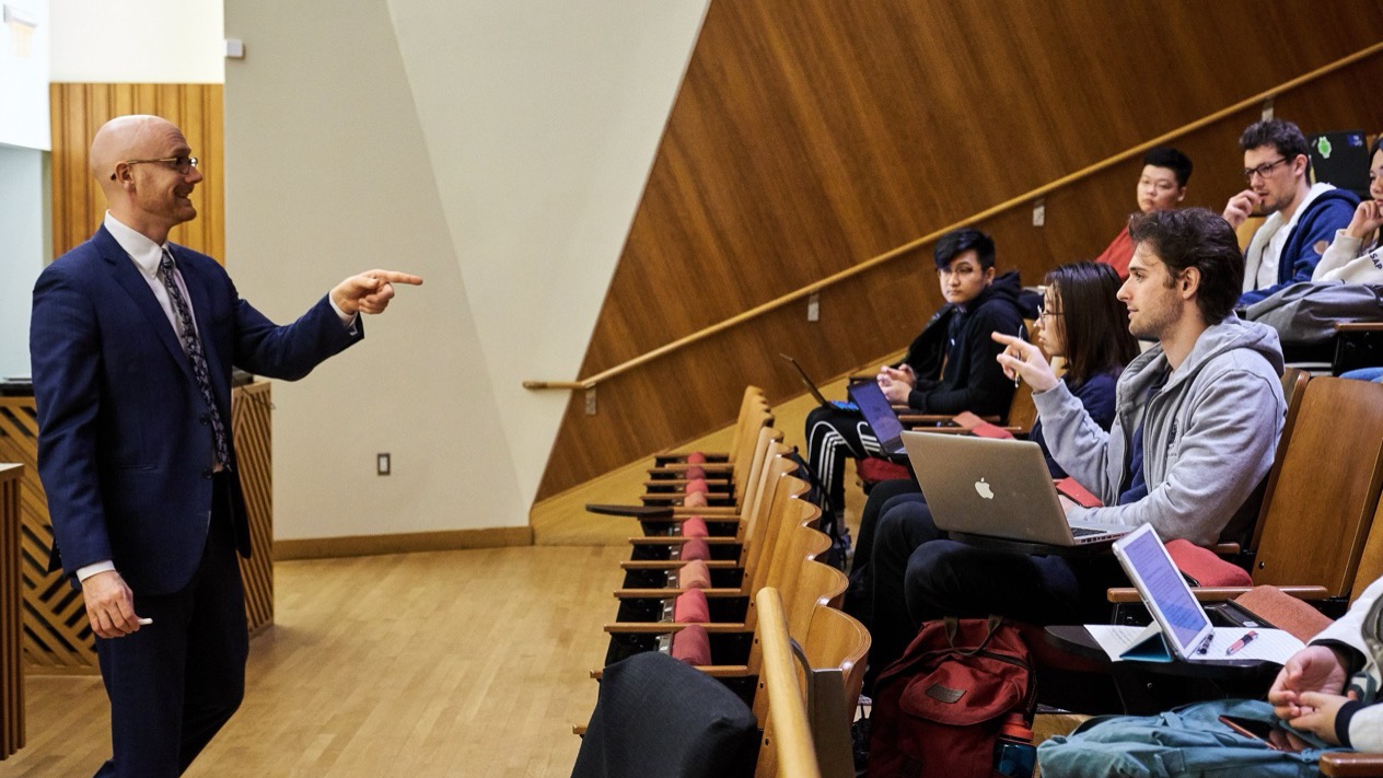 Professor in navy suit and glasses teaching in a tiered wooden lecture hall, gesturing while addressing approximately 15-20 students seated in elevated rows.