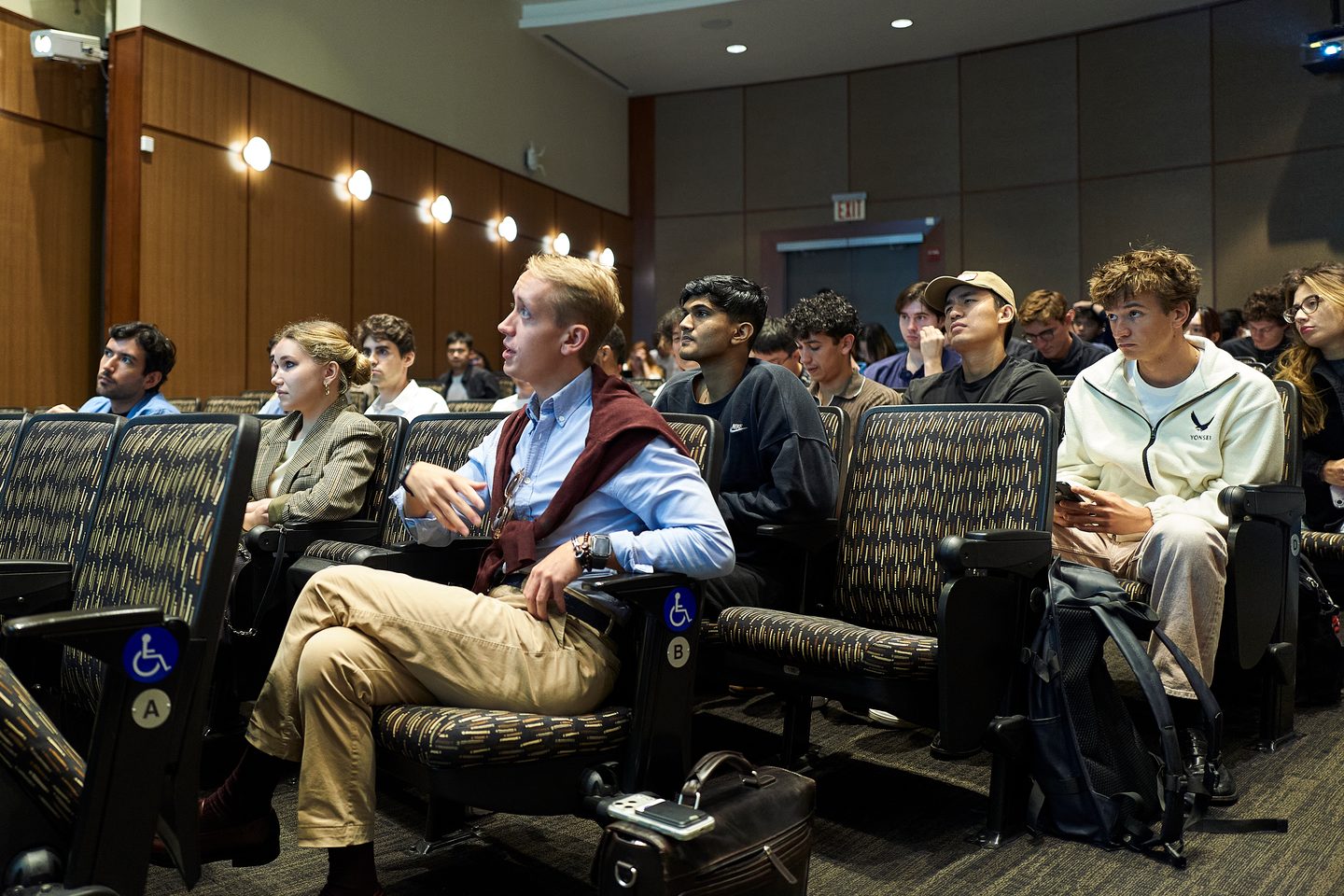 Attendees seated in an institutional room with accessibility-designated seating, watching or waiting during what appears to be a formal proceeding or meeting.
