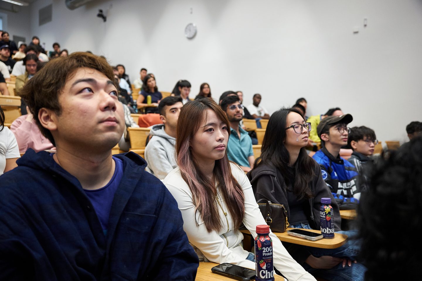 Students attending a lecture in a university lecture hall, seated at individual desks and looking upward.