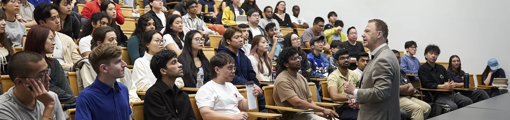 A session in a large tiered lecture hall, with a speaker in a gray blazer addressing rows of students who are seated and listening attentively.
